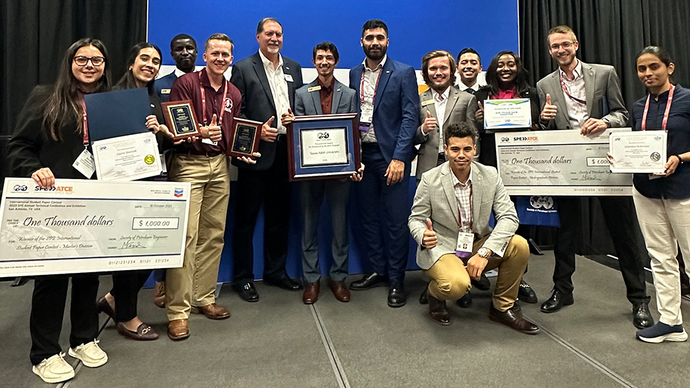 thirteen people dressed in nice attire stand on a stage while holding certificates, framed awards and large posters with award amounts on them 