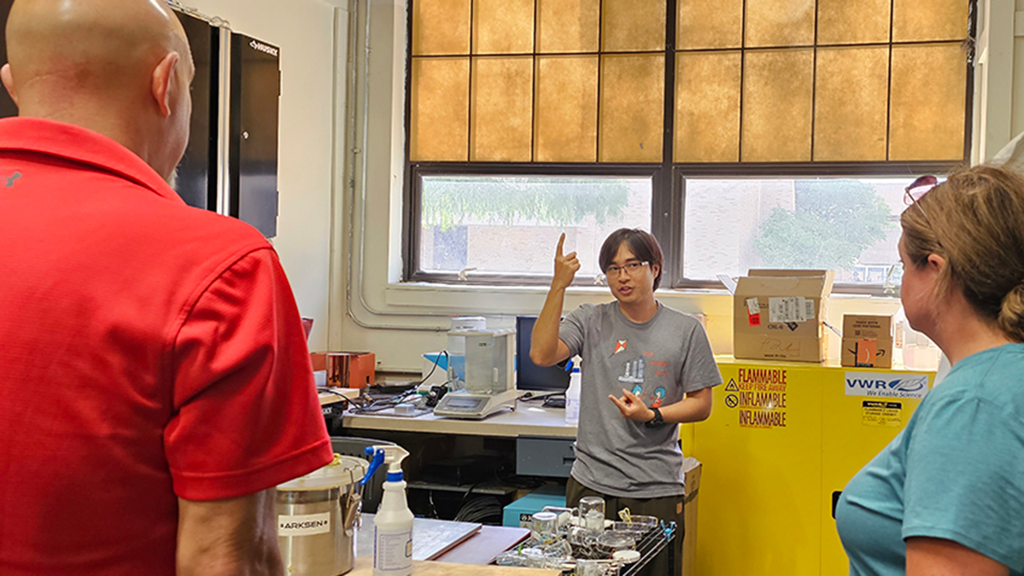 Two people stand looking at another man in a gray shirt gesturing about equipment inside an older warehouse-type building. 