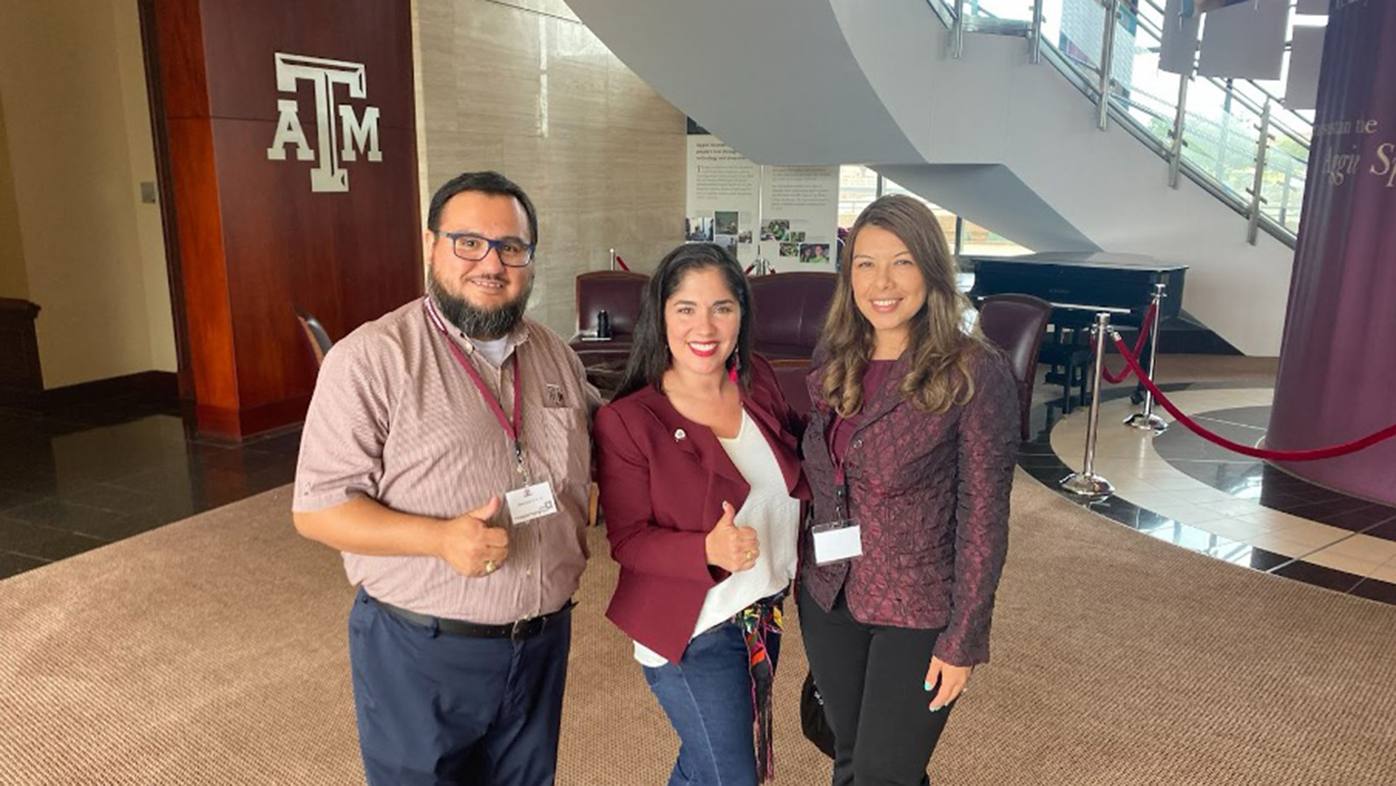 Dr. Jennifer Carvajal in an academic building with Mario Hernandez and Tamara Cuellar, two leaders at a former student association, who are giving a thumbs up with the A&M logo in the back. 