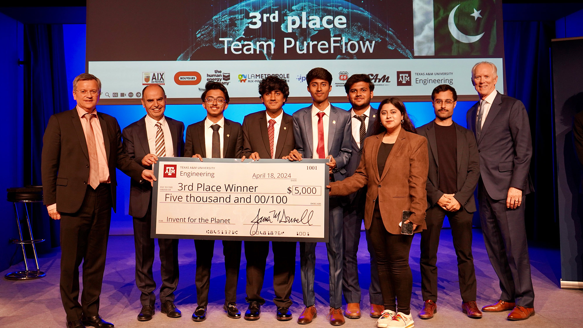 Nine people stand on a stage by a large check for $5,000.