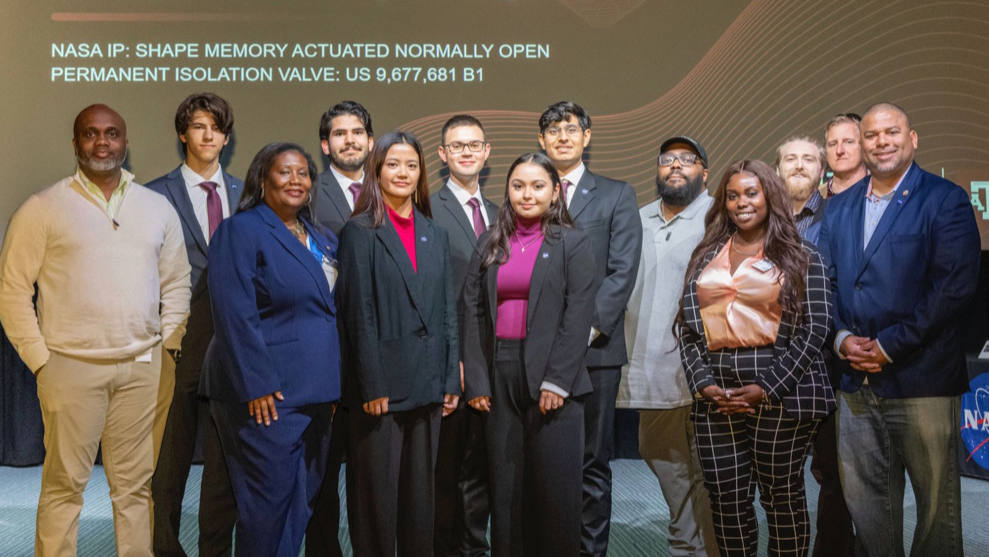 A group of people standing on a stage. In the background is a PowerPoint slide that reads “NASA IP: SHAPE OF MEMORY ACTUATED NORMALLY OPEN PERMANENT ISOLATION VALVE: US 9,677,681 B1.”