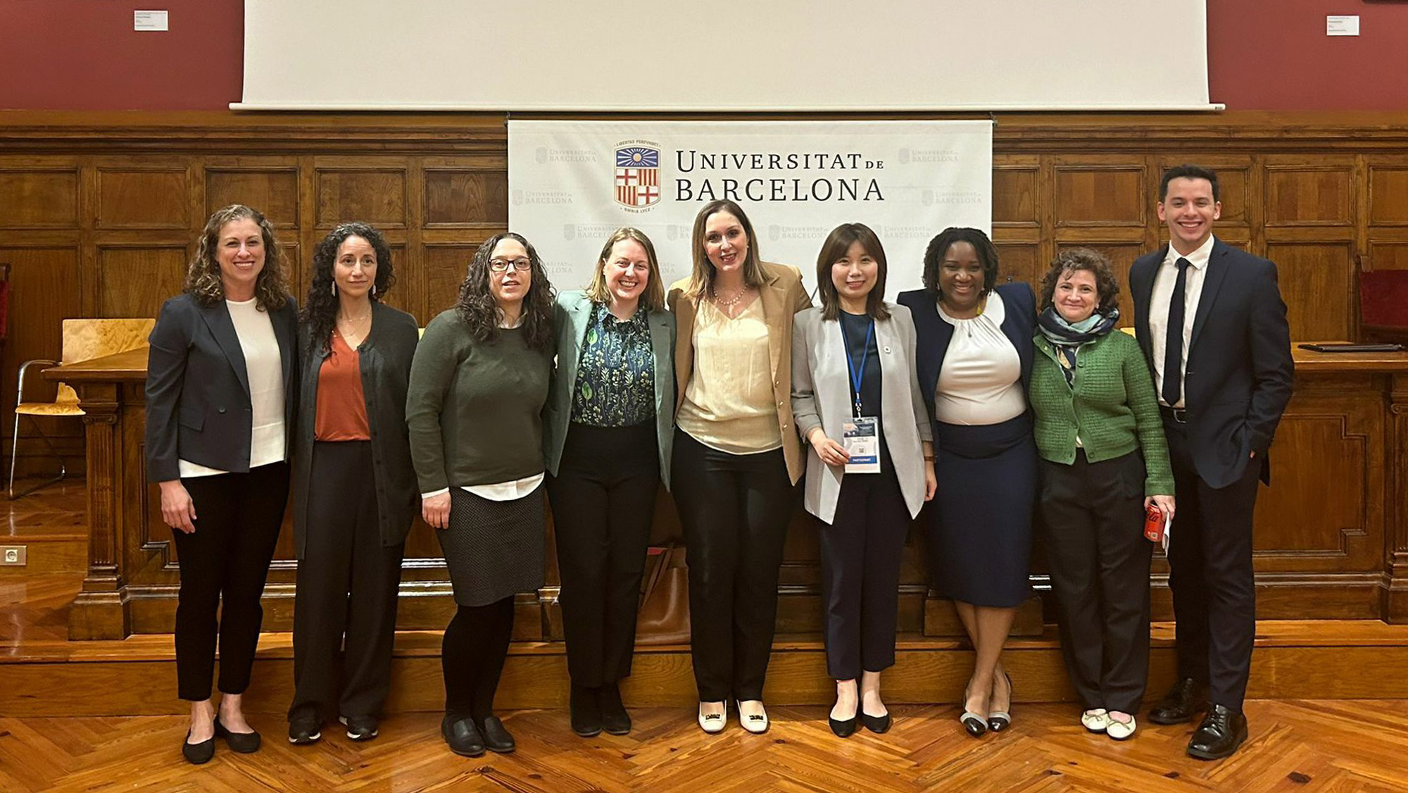 A group of eight women and one man in front of a banner for the University of Barcelona.