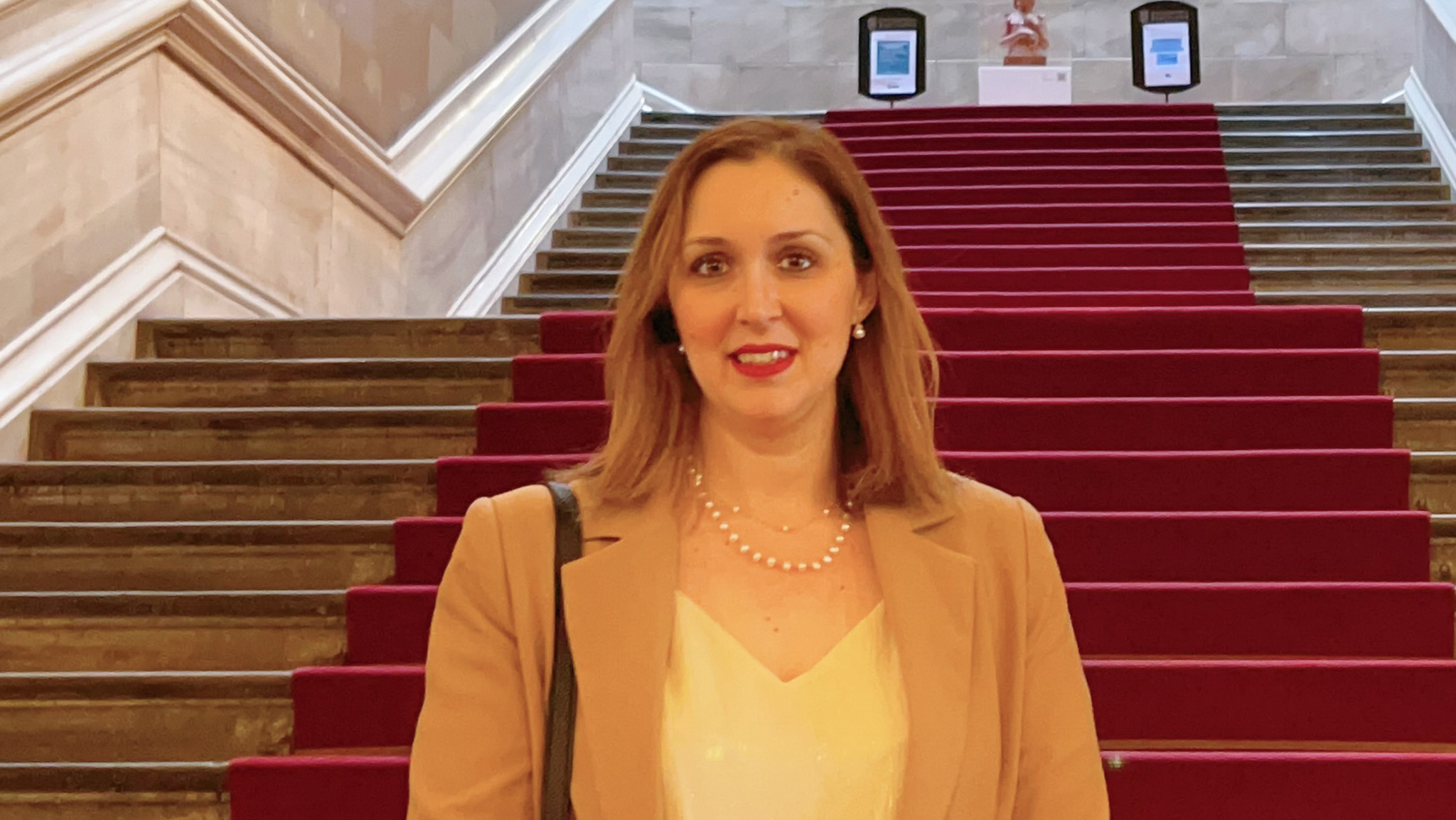 A woman stands on the grand stairway of a historic building.