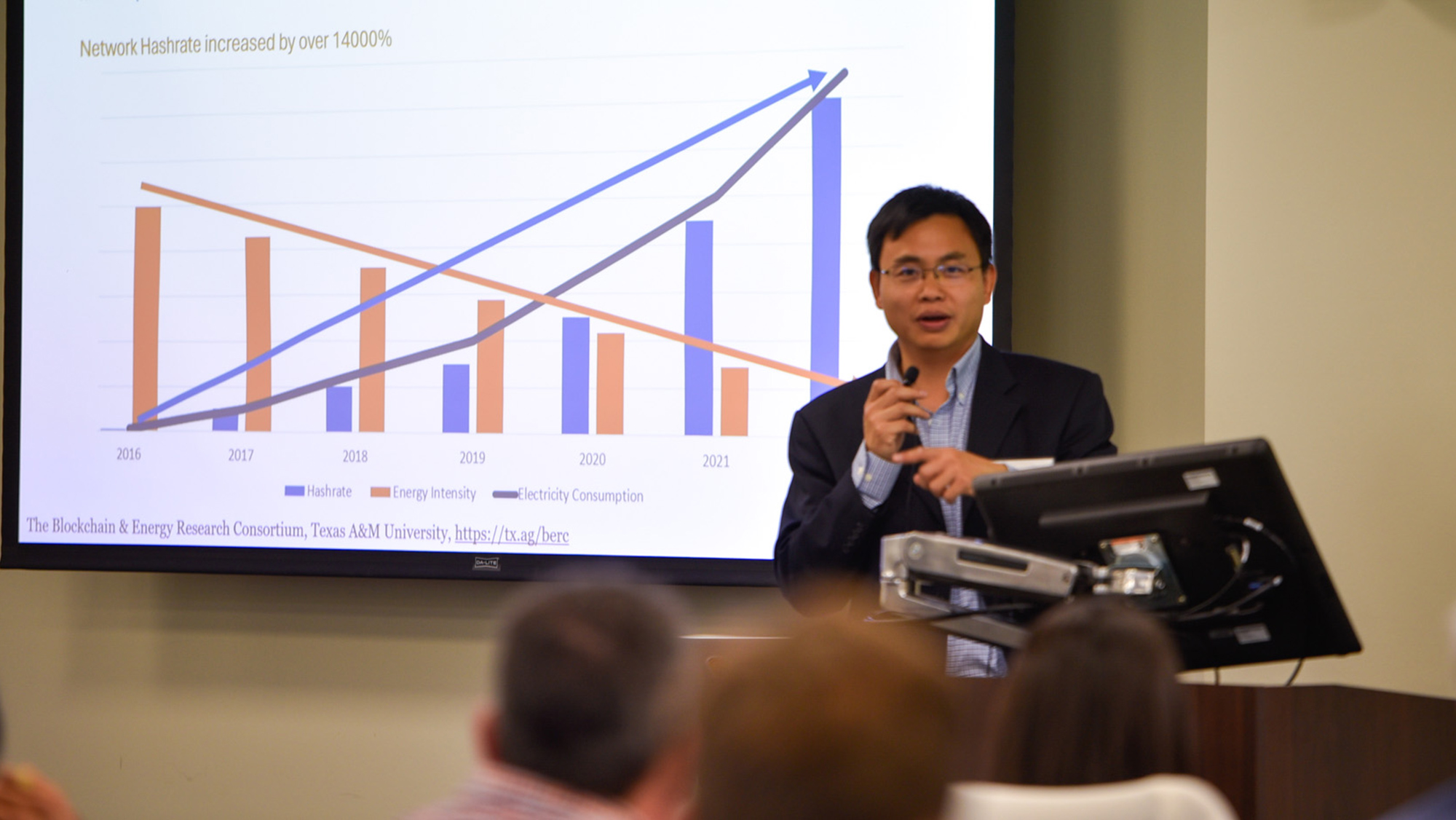 A person presenting a slide on Mining Energy Intensity and Hashrate to an audience in a conference room setting.