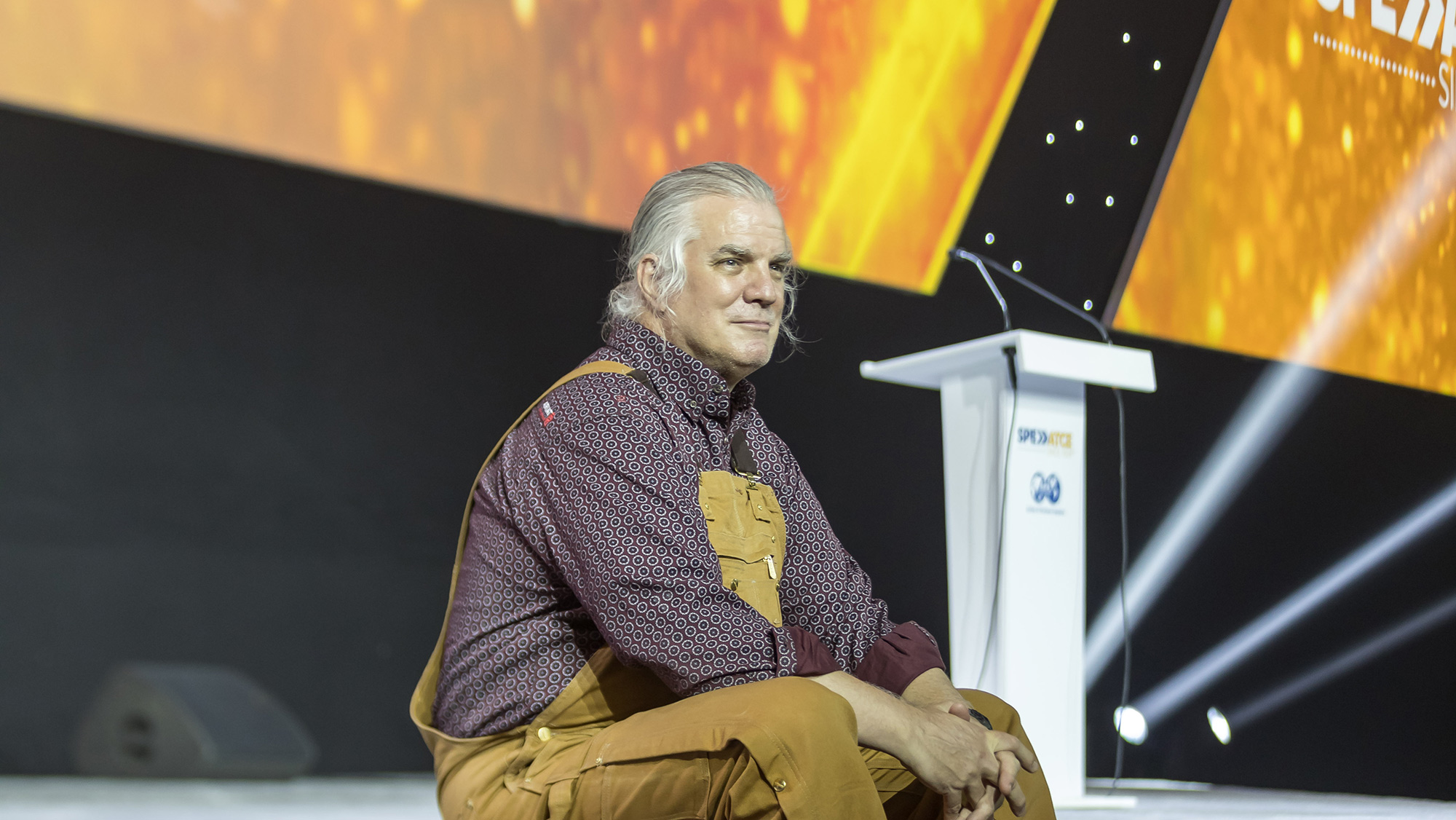 A man with grey hair wearing tan overalls sitting on the edge of a stage.