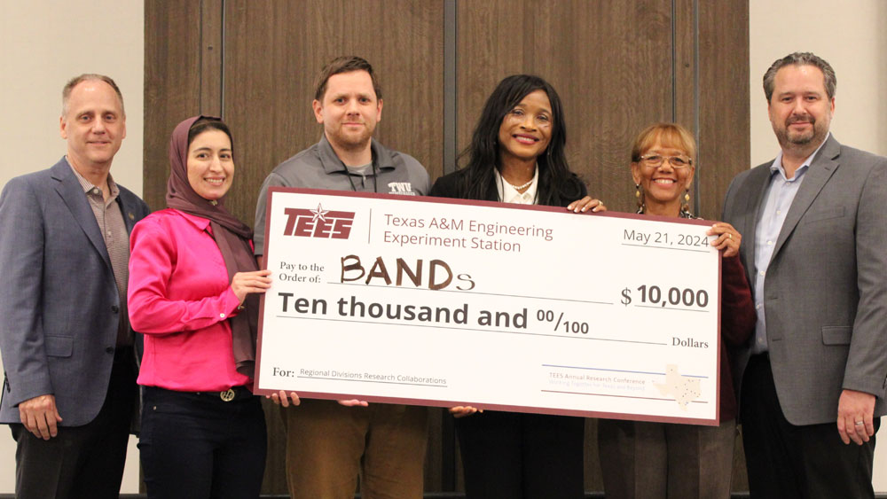 Three men and three women stand on stage holding a giant check. 