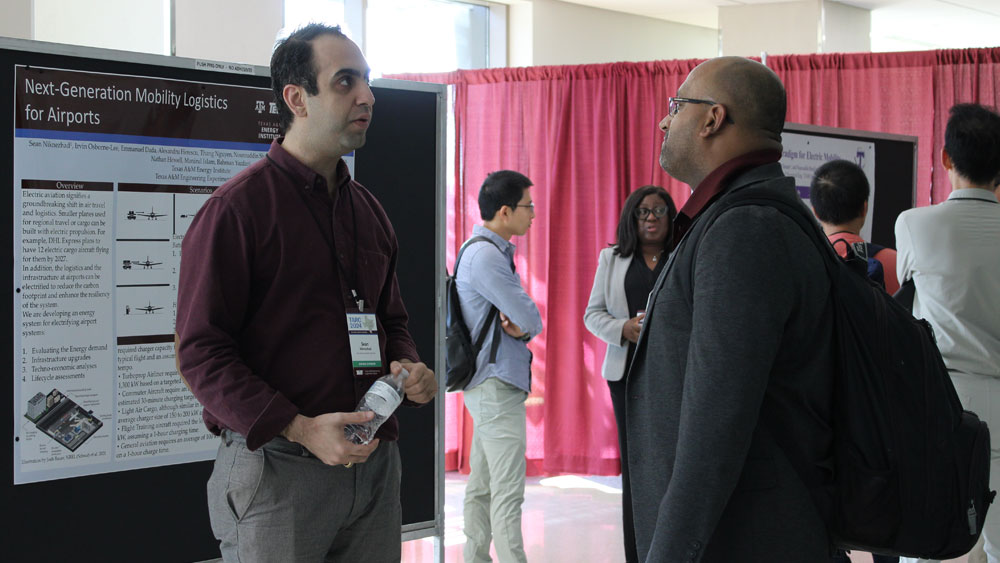 Two men stand talking in front of poster display board. 