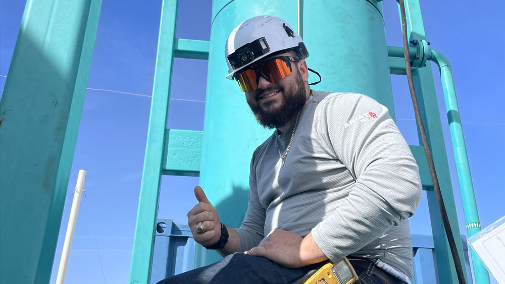 Man in jeans, long-sleeve t-shirt, hardhat and goggles sits on machinery. 