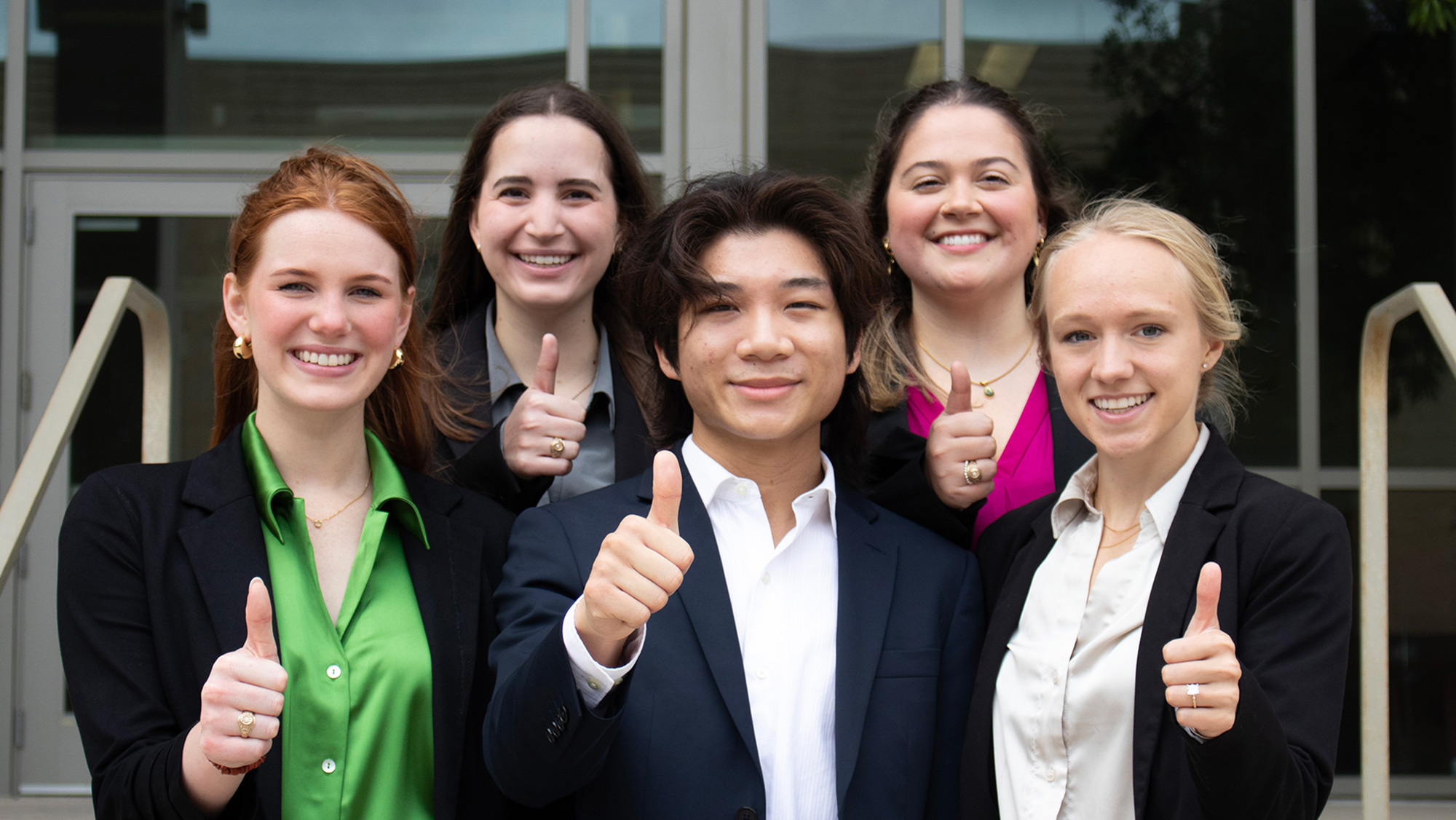 Six students dressed in professional attire with their thumbs up on the steps of a campus building.