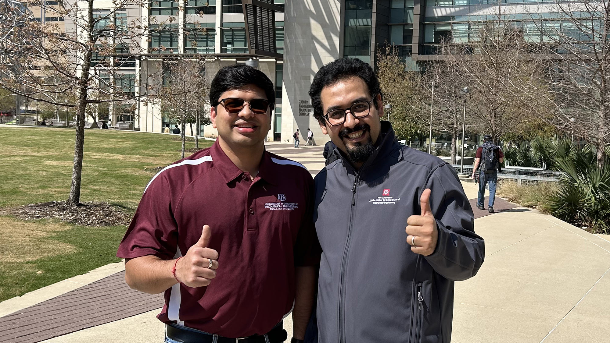 Two people standing and smiling with thumbs up in front of a building.