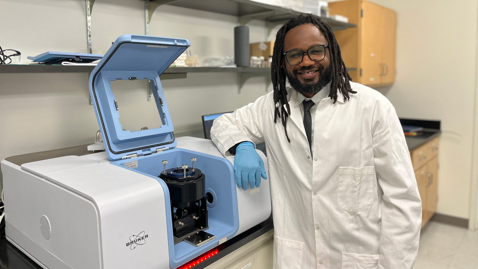 Scientist in lab coat and gloves standing beside an open laboratory equipment in a research lab.