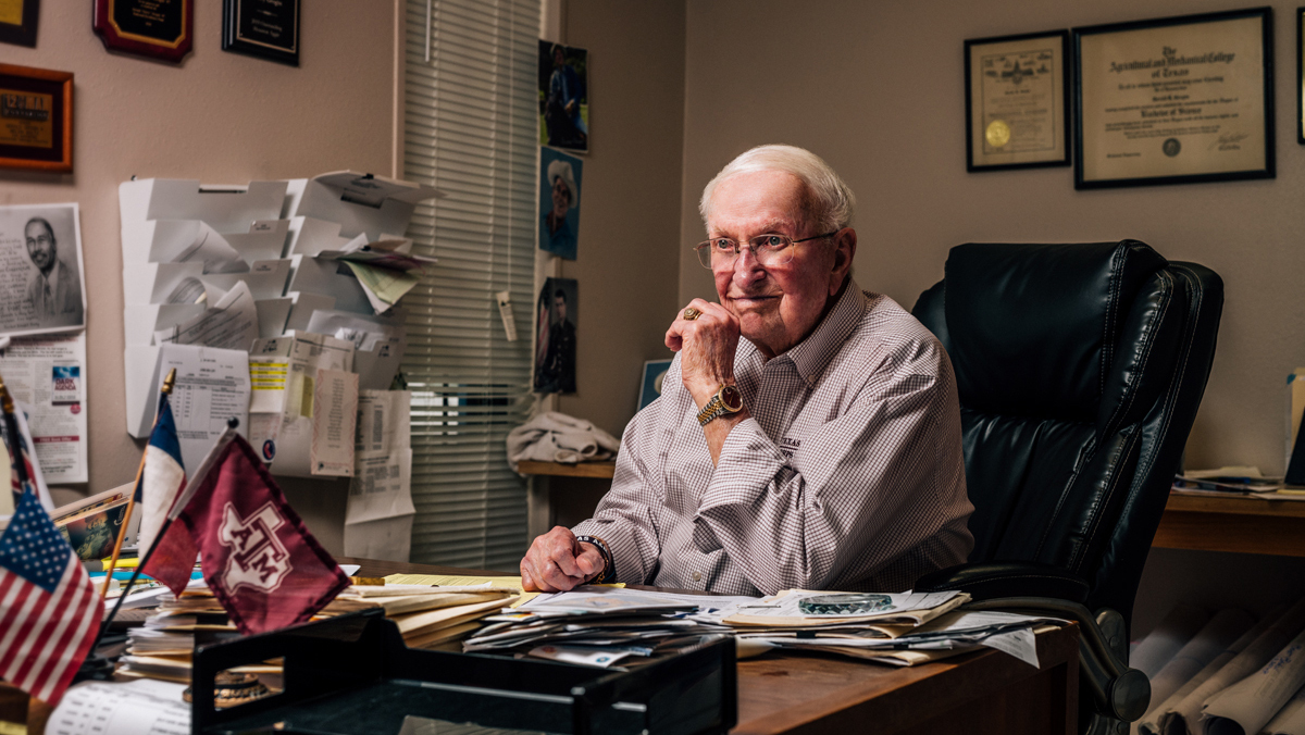 Man sitting at a desk with papers and personal mementos, with framed diplomas and certificates on the wall behind him.