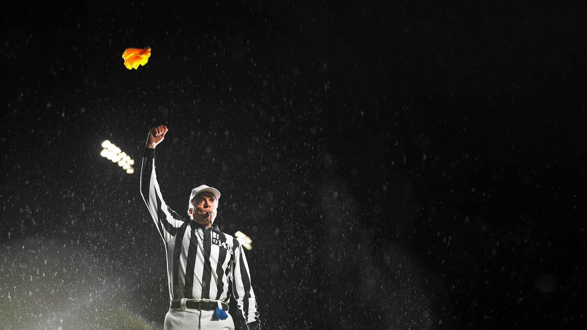 A referee throwing a yellow flag and blowing a whistle on a football field at night. 