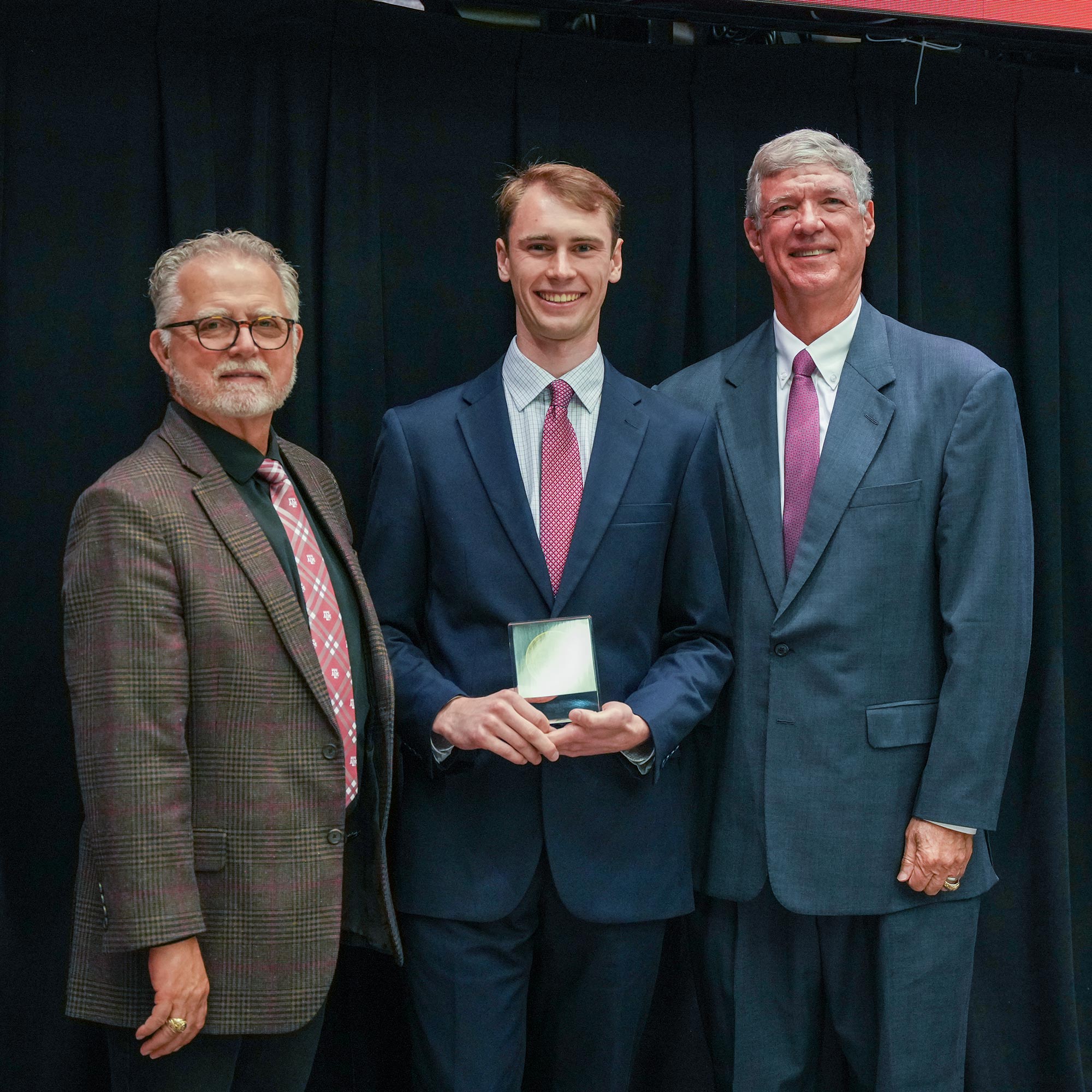 Three men standing, one with an award.