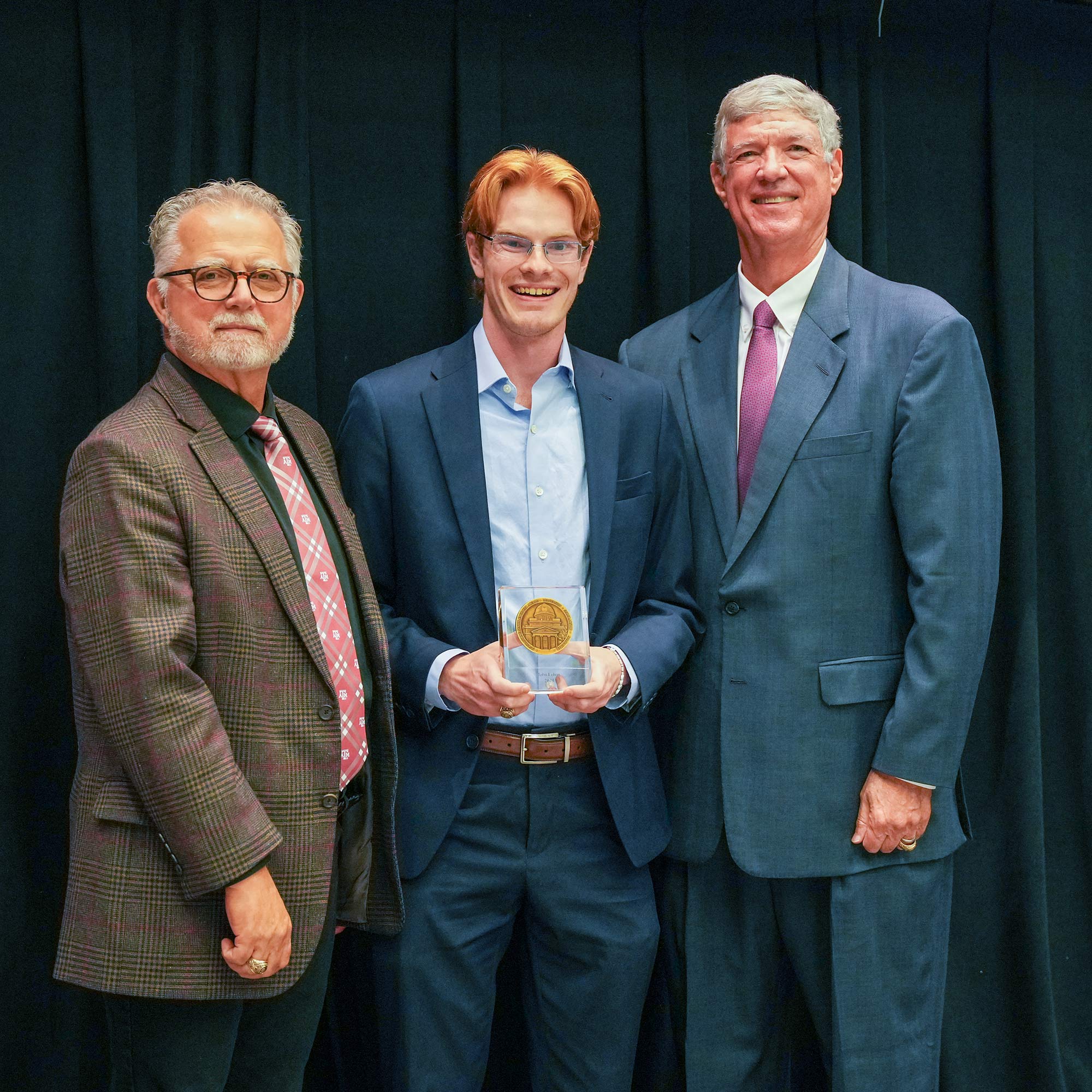 Three men standing, one with an award.