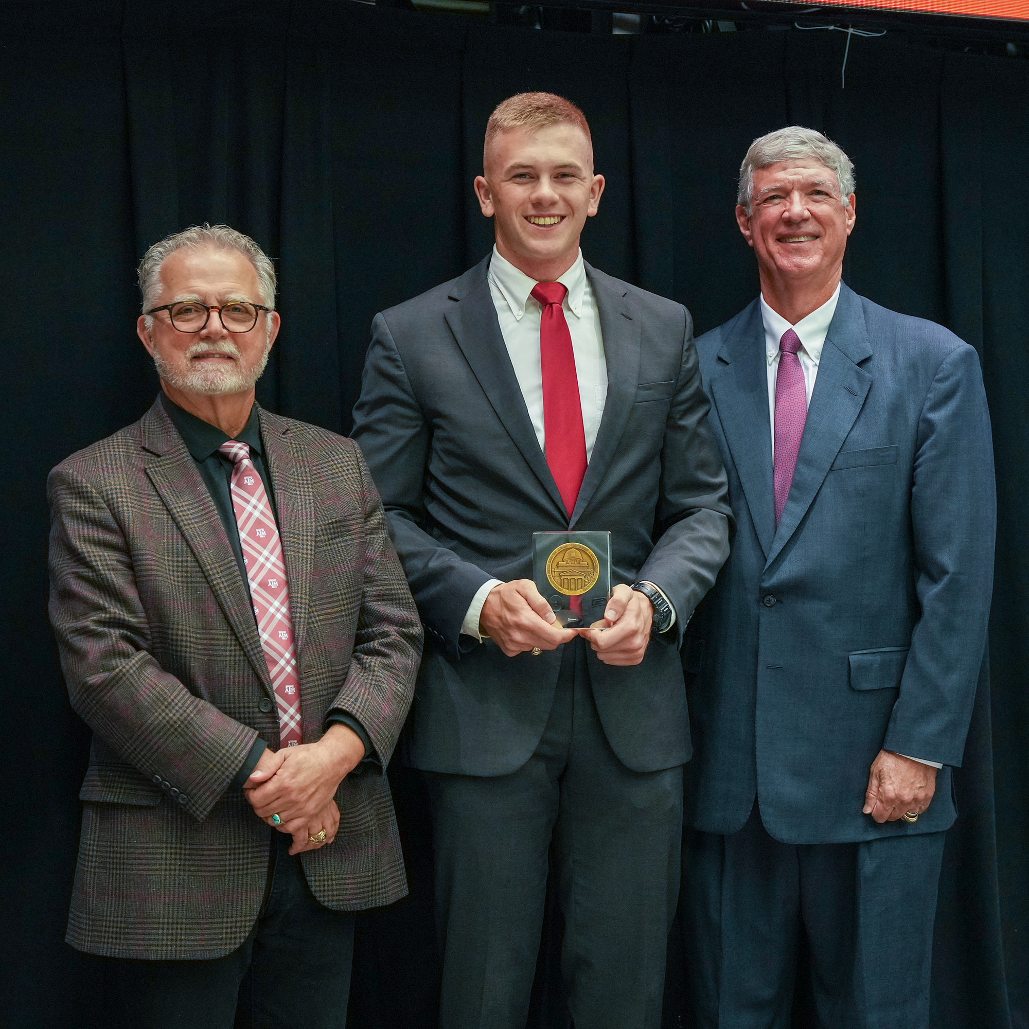 Three men standing, one with an award.