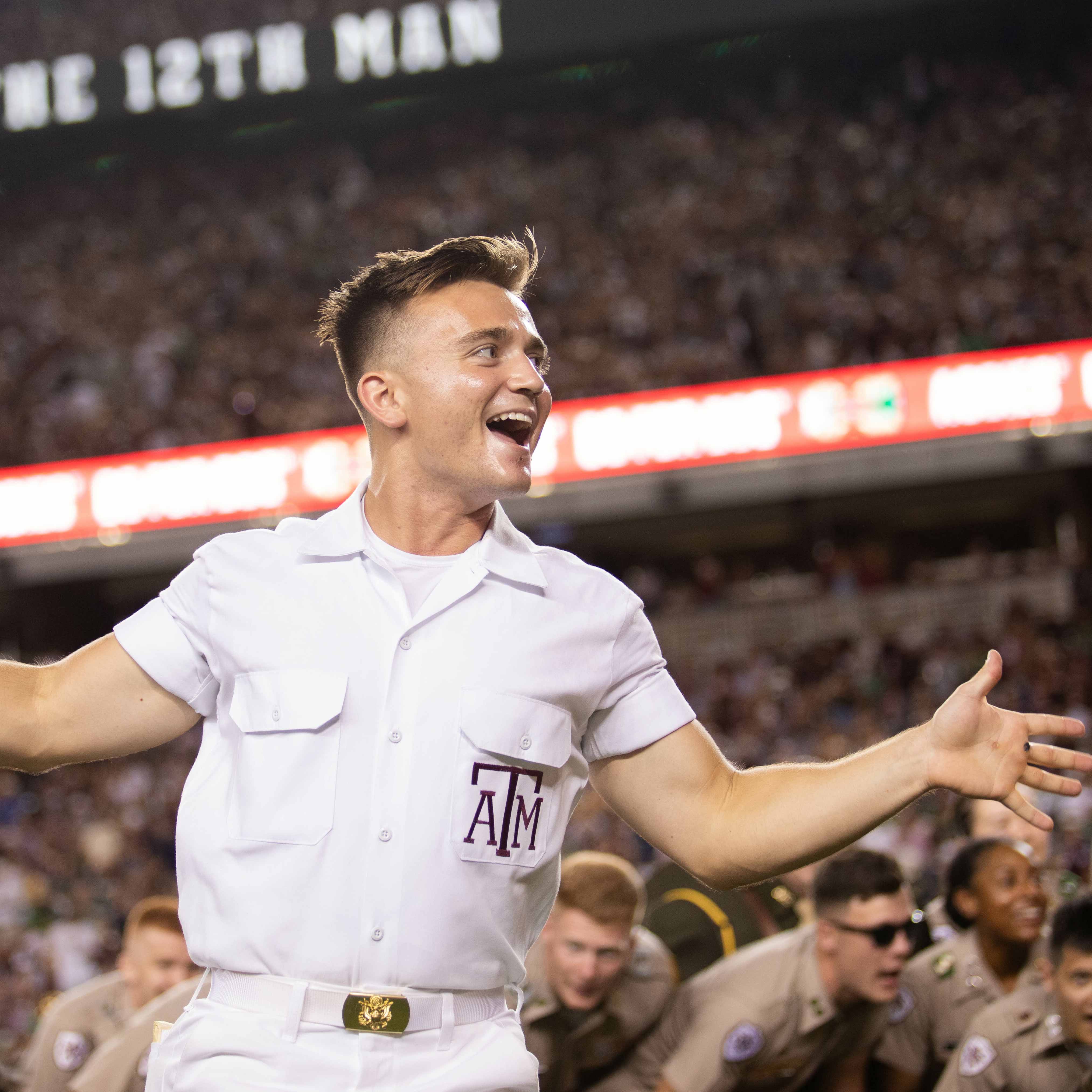 Texas A&amp;M yell leader wearing a white uniform performing a yell with a large crowd in the background.