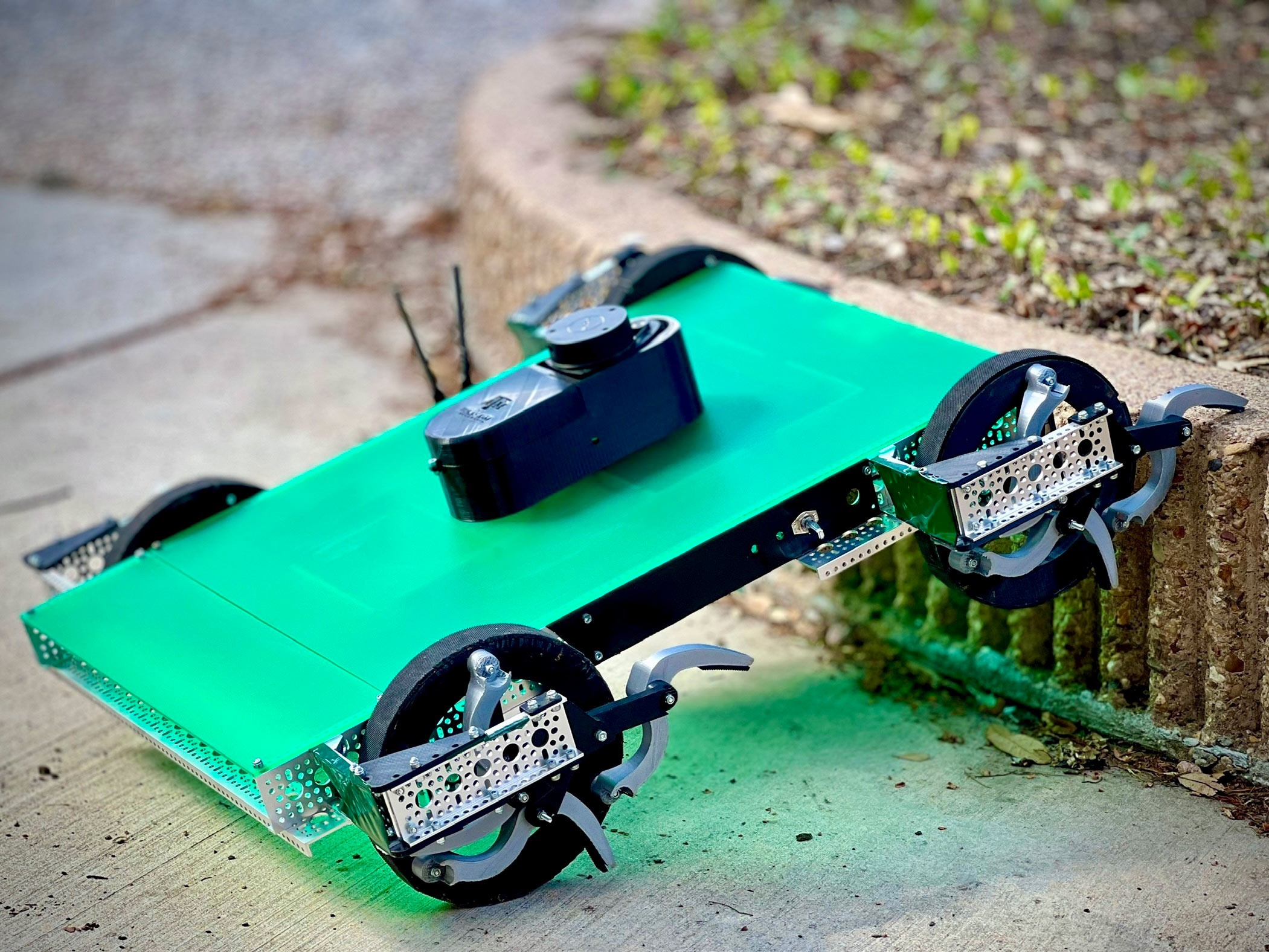 The wheel of a green and black robot extends a claw to grab a short, concrete barrier. 