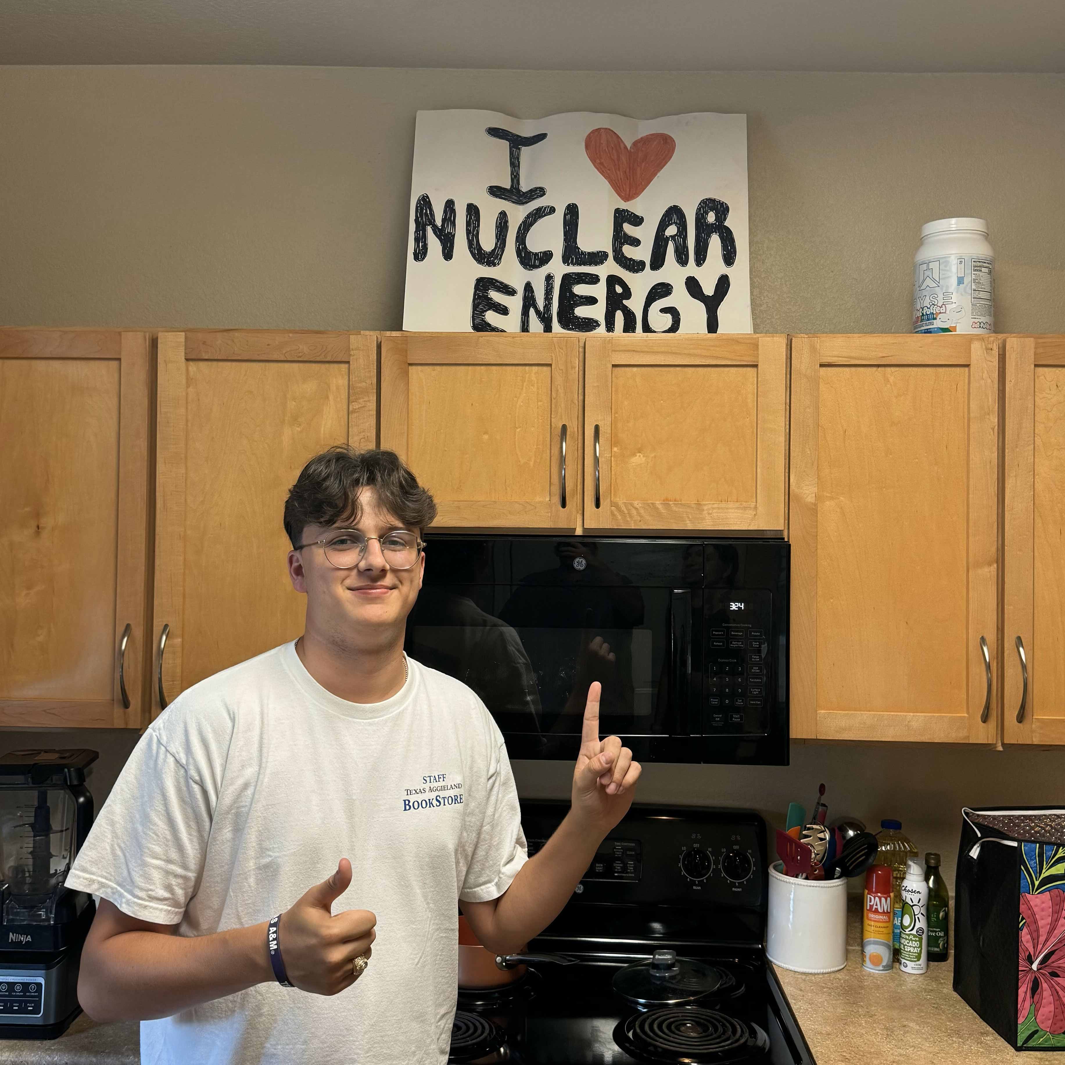 A man in a t-shirt stands in a kitchen, smiling and pointing toward a sign that reads I heart nuclear energy.