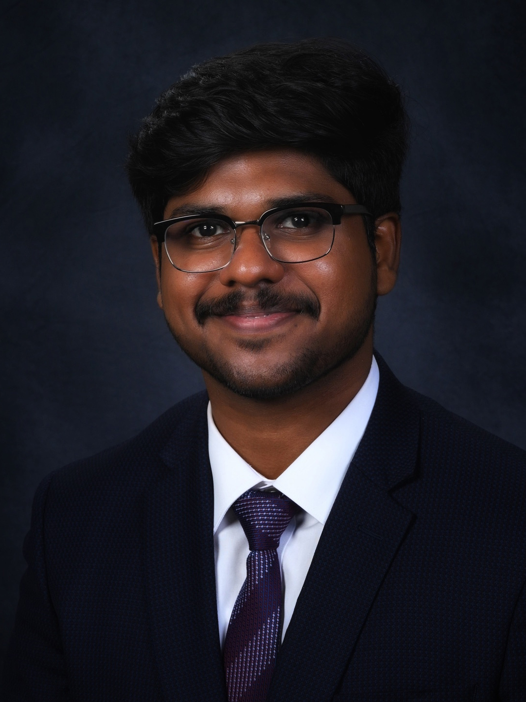 Headshot of a young man wearing a black suit and glasses smiling at the camera.