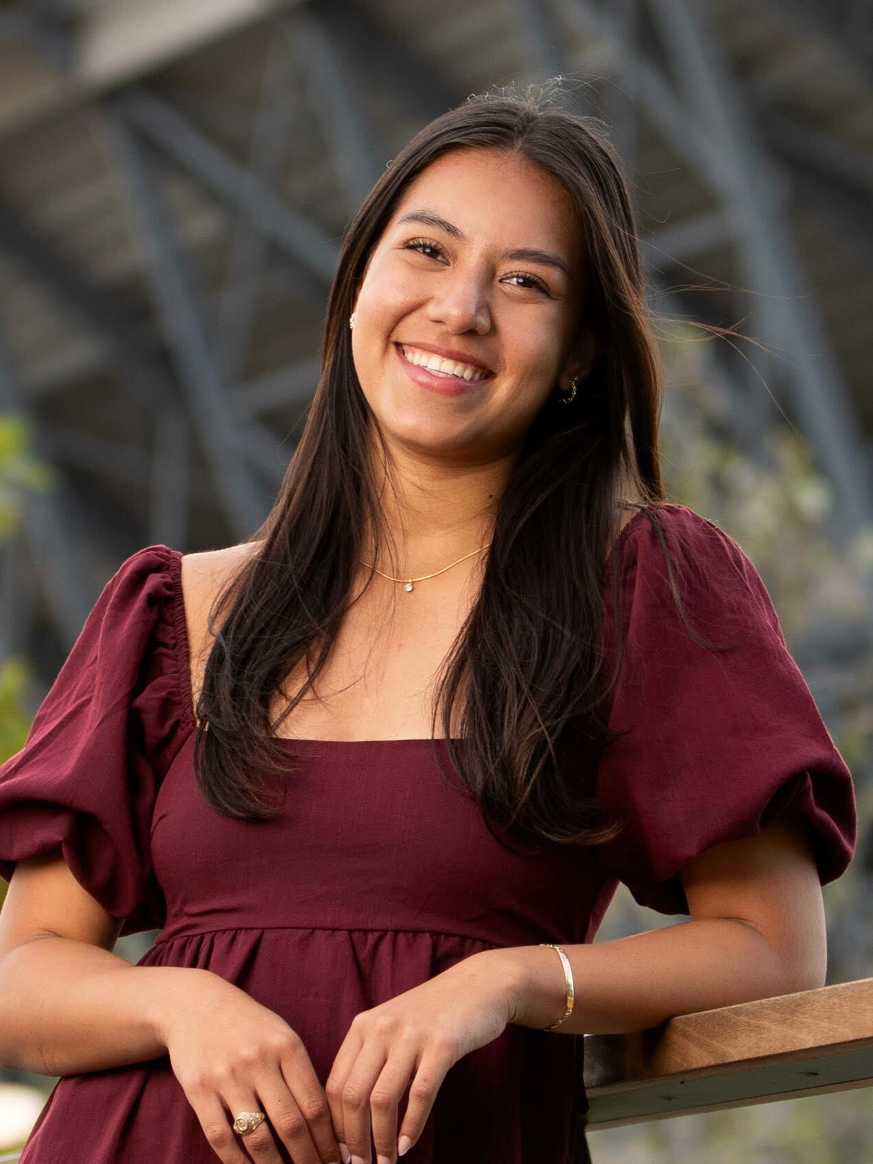 A young woman smiles while leaning on a wooden railing, wearing a burgundy dress.