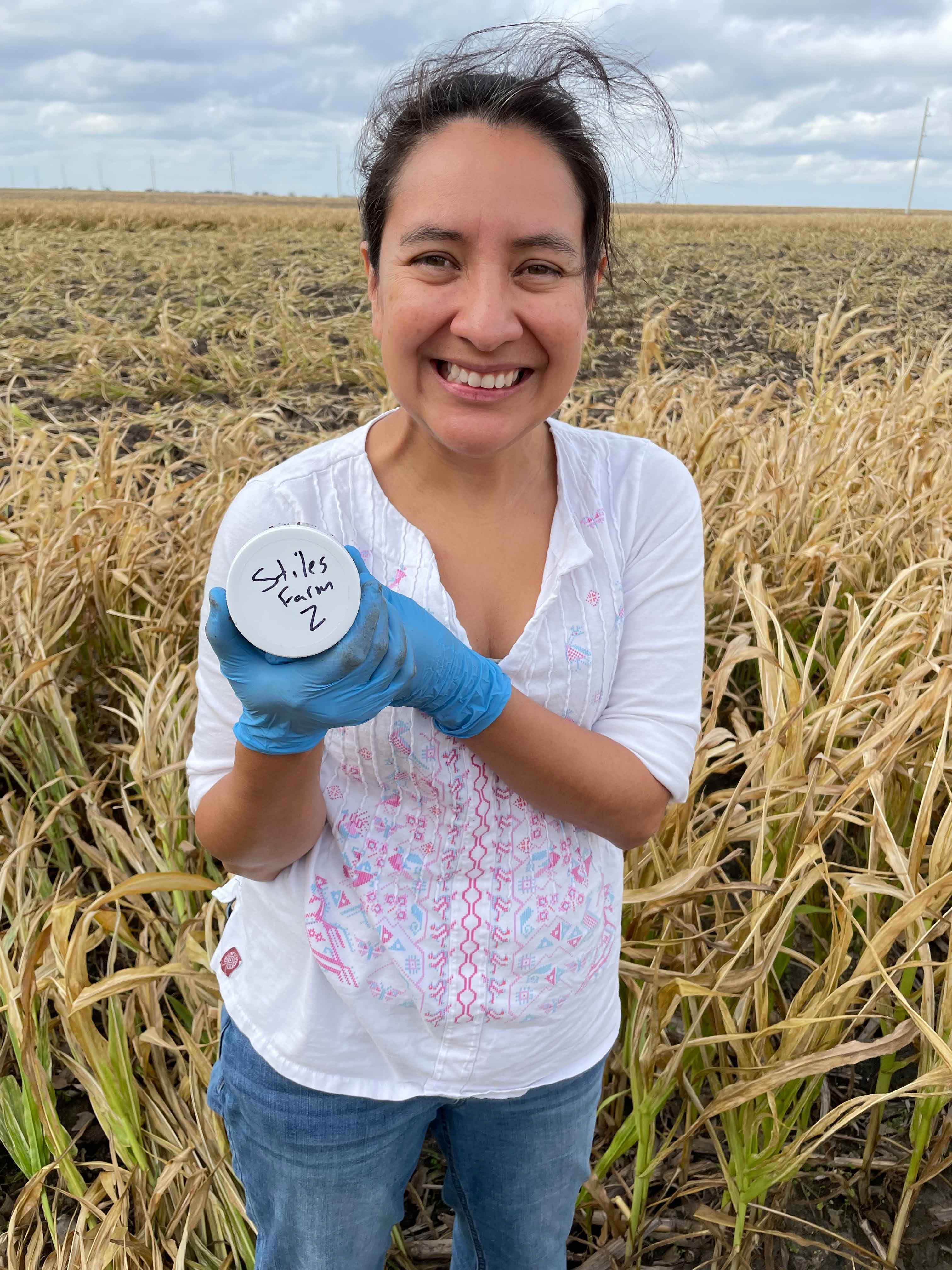 A woman stands in a crop field holding a sample container.