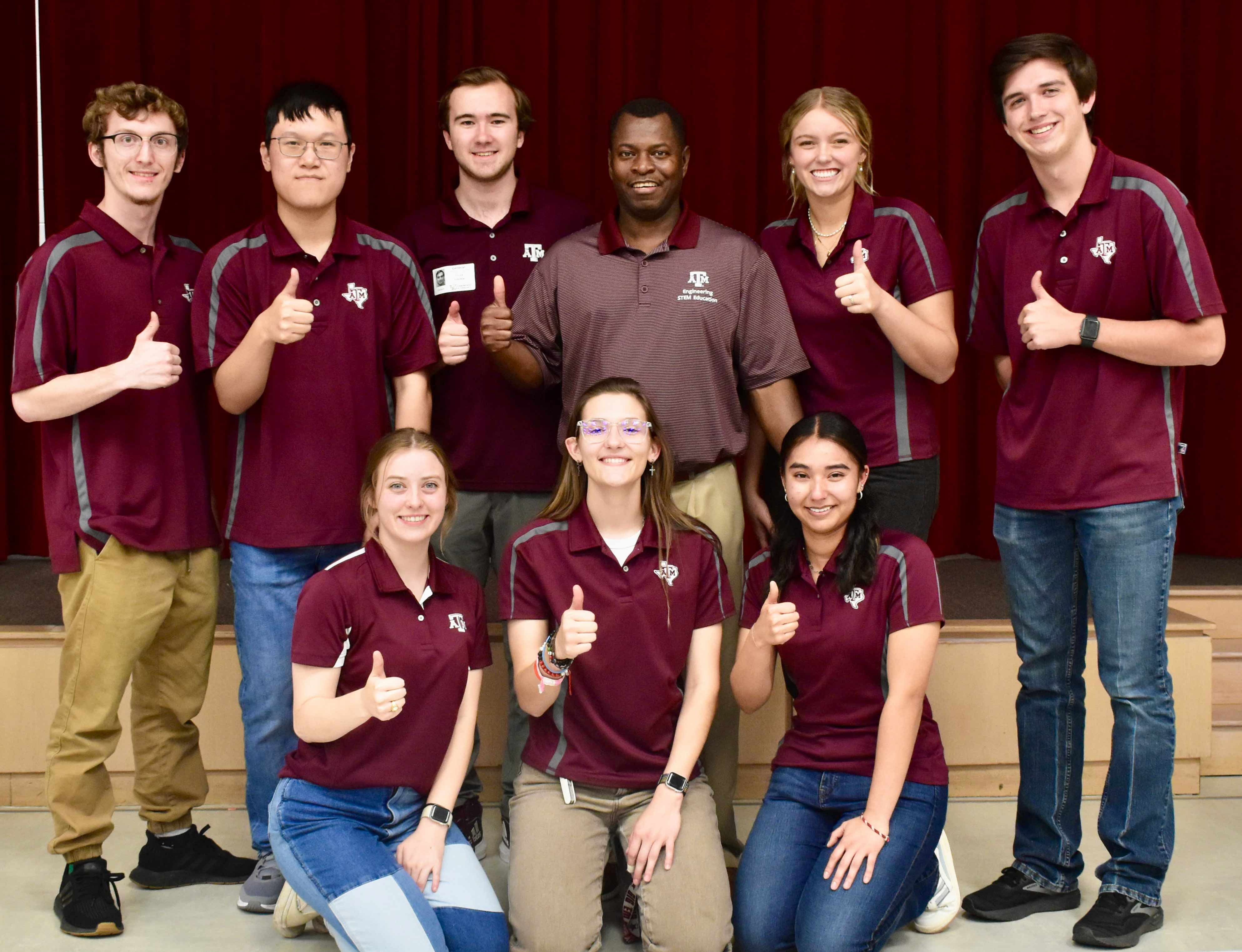 A professor and a group of eight students smile while giving thumbs up.