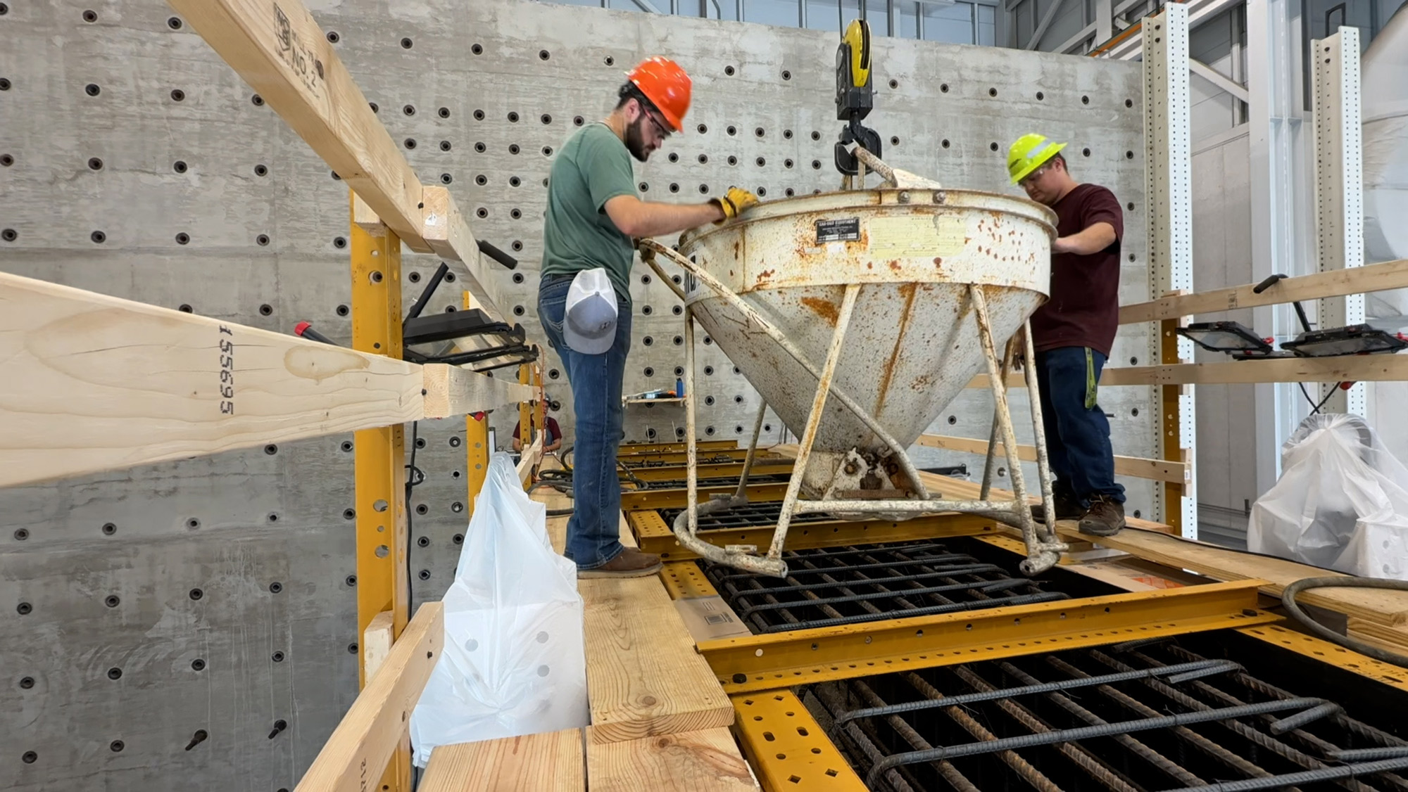 Two workers move a crane-lifted cement bucket over a large structural framework.