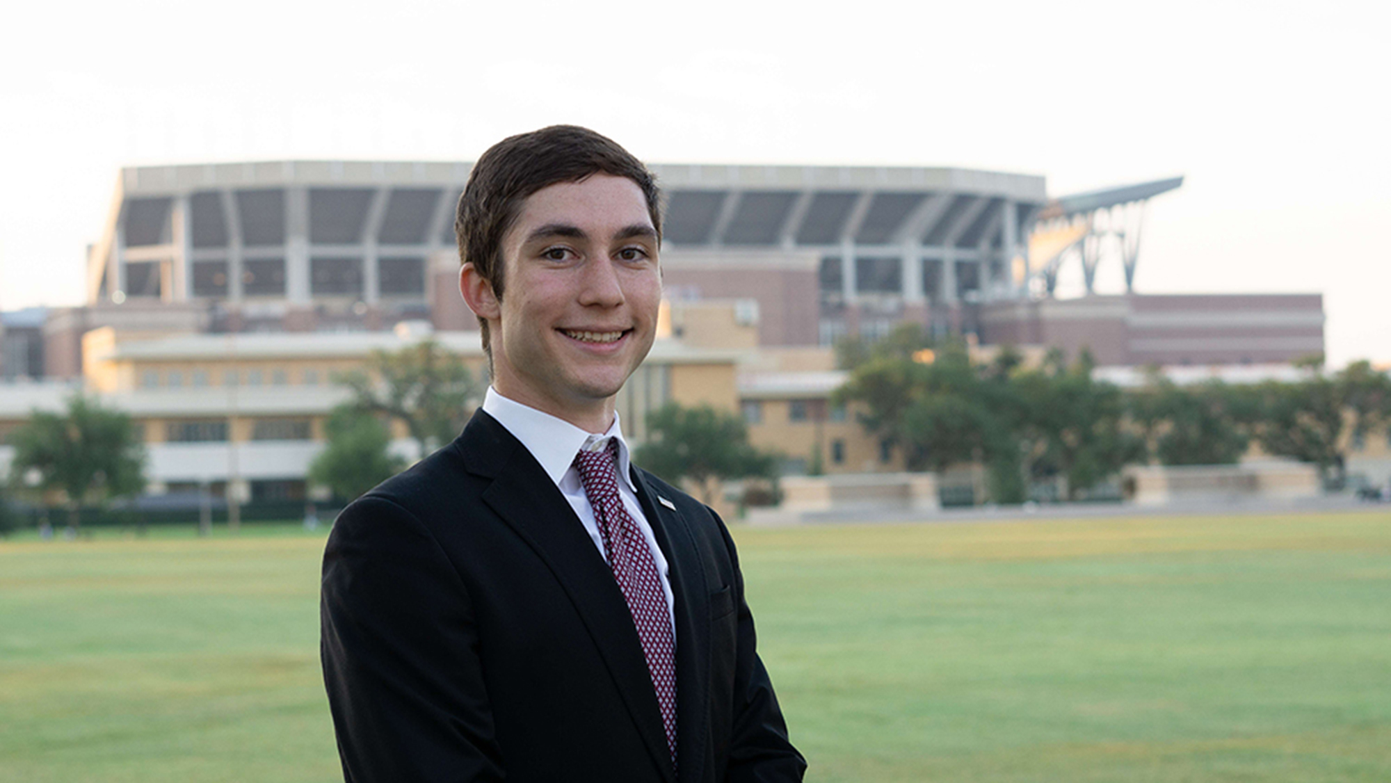 A young male wearing a suit jacket and tie stands smiling at the camera with a green field and football stadium in the background.  