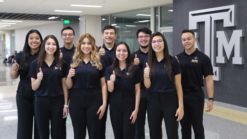 Group of students standing next to A&M logo with thumbs up.