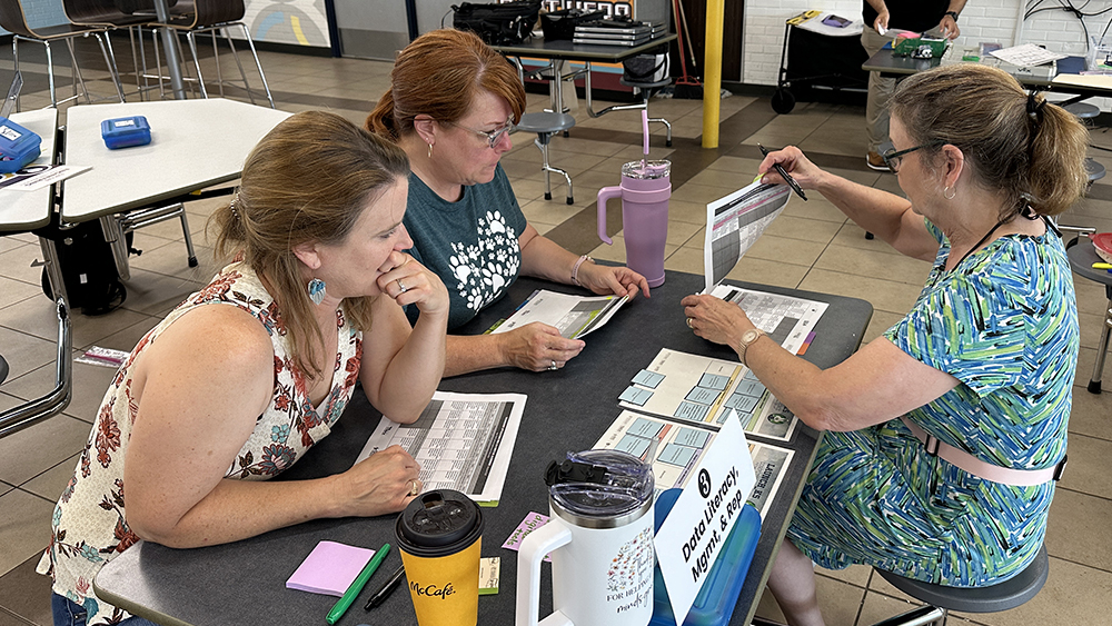 Three teachers sit around cafeteria table looking at lesson plans.