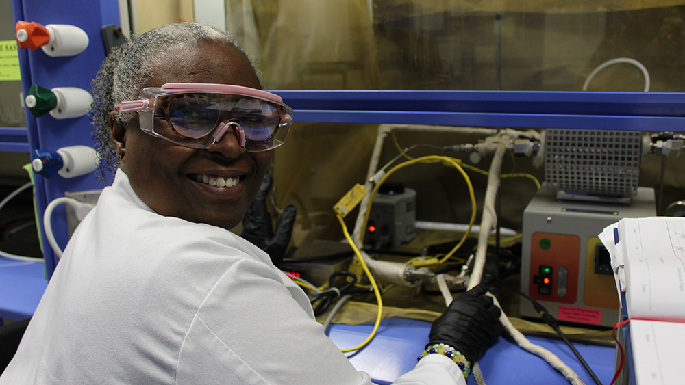 Woman wearing safety goggles smiles at the camera while holding on to lab equipment.