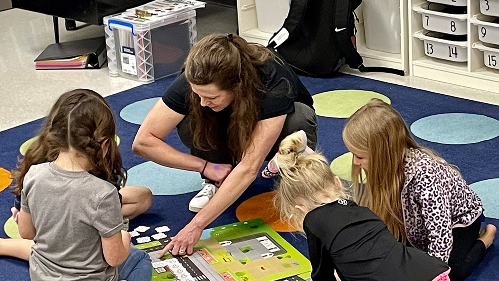 A woman works with elementary-age students to program small robots to move along a map. 