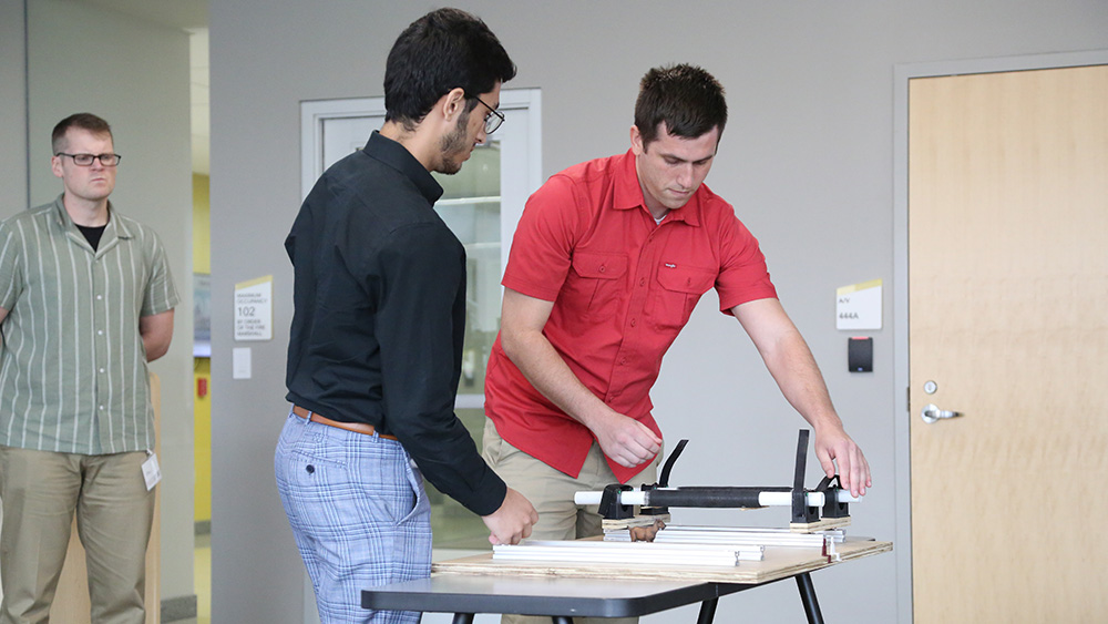 Two students adjust a simple mechanical mechanism on a tabletop while a third person observes.